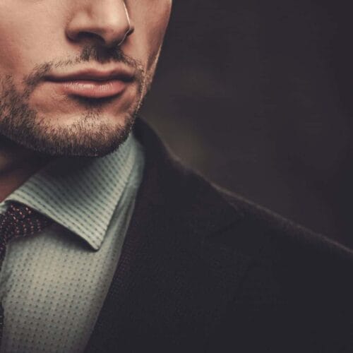 Close-up of the lower half of a man's face, showcasing a well-groomed beard and mustache that exhibit masculine qualities. He is wearing a blue dress shirt with a small checkered pattern, a patterned tie, and a dark blazer. The background is blurred, highlighting the subject's manly traits women find attractive.