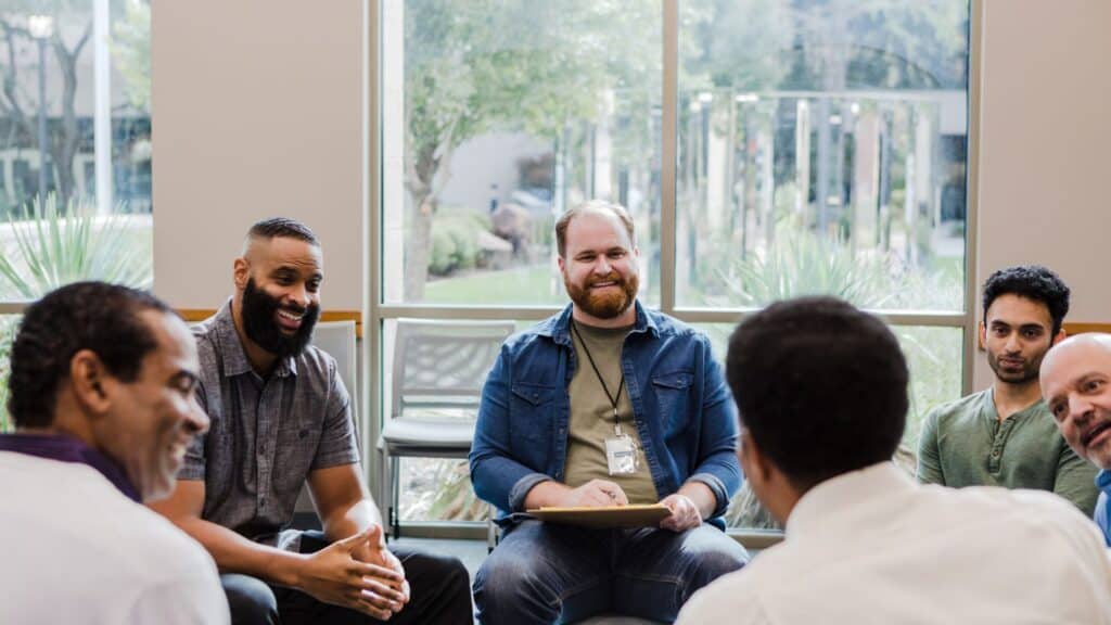Home 4 A group of men, including Santosh Acharya, a Family Leadership Coach, sit in a circle engaged in conversation. They appear to be in a casual meeting or discussion in a room with large windows showcasing greenery outside. The men are dressed casually, and one holds a clipboard.