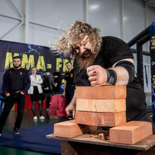 At a martial arts event, a man with long hair and a beard channels unexpected power as he attempts to break a stack of clay bricks with his hand. Spectators and officials stand nearby, observing the action, captivated by this display of strength and vulnerability.