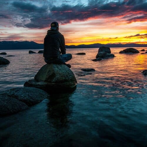 A man sits on a rock by a tranquil lake, watching a vibrant sunset with orange, pink, and purple hues in the sky. The calm water reflects the colorful sky, and distant mountains frame the horizon.
