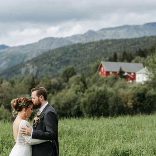 A bride and groom, embodying the essence of a good marriage, embrace in a lush green field with mountains in the background. A red and white house sits among trees under a cloudy sky, symbolizing the foundation of their promising future together.