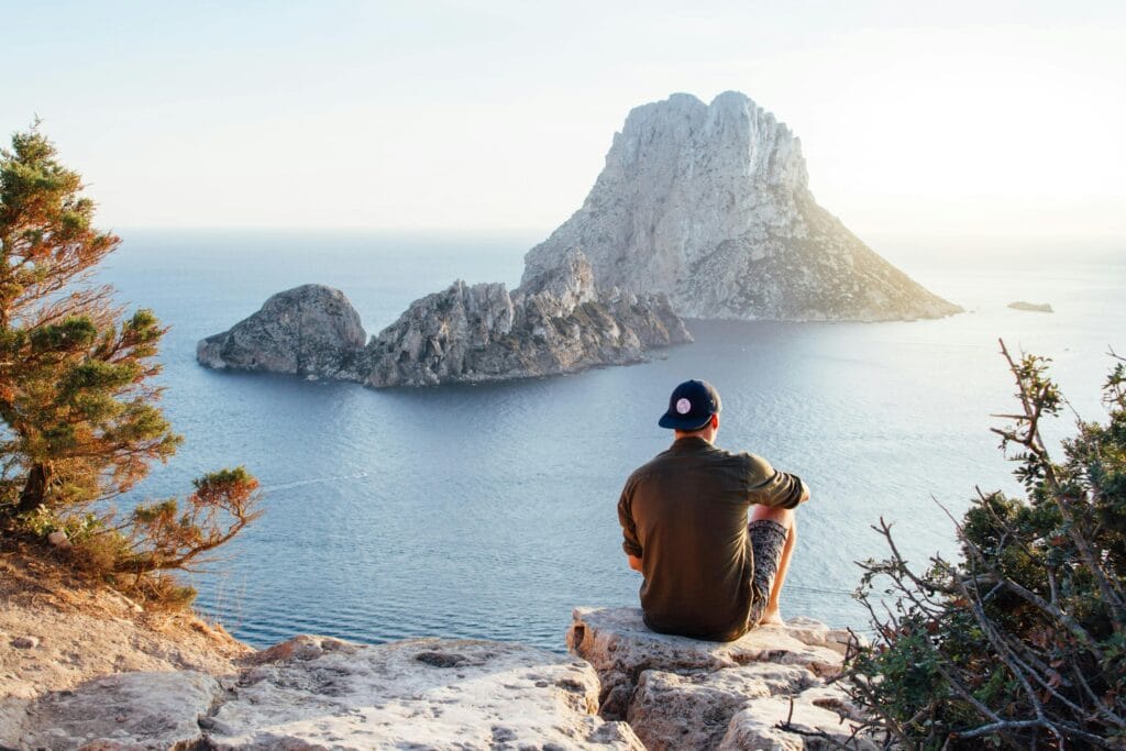 Why Men's Issues? 1 A person in a cap, contemplating men's issues, sits on a rocky cliff overlooking a calm sea with a large rock formation in the distance. Green shrubs and trees surround the scene under a clear sky, offering peaceful solitude.