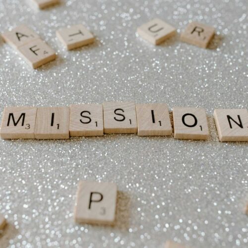 Scrabble tiles spelling "MISSION" are arranged on a sparkling, glittery surface, surrounded by other scattered tiles. It's as if they whisper: Stop Setting Goals like men setting sparks—start setting fires to ignite your true purpose.