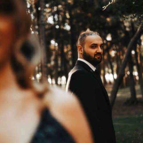 A man in a suit stands in focus against a blurred forest background, seemingly avoiding conversation. In the foreground, the blurry outline of a woman's shoulder and hair is visible. The scene has a warm, golden glow, hinting at an important relationship discussion just out of reach.